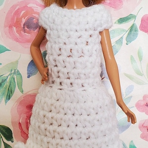 Puede incluir: Un vestido blanco de ganchillo con un dobladillo con volantes, diseñado para una muñeca.