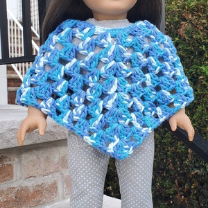 May include: A doll wearing a blue and white crocheted poncho, grey pants with white polka dots, and floral print shoes.