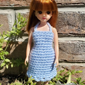 May include: A doll with red hair wearing a light blue crocheted dress. The doll is standing in front of a brick wall and surrounded by green plants. The dress has a halter top design.