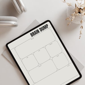 Può includere: Un tablet digitale con bordo nero mostra un modello "BRAIN DUMP". Il modello include sezioni per gratitudine, casa, lavoro, progetti, personale, altri e note. Sono visibili anche delle cuffie argentate e un vaso con piante essiccate.
