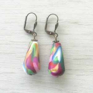 Peut inclure: Une paire de boucles d'oreilles en forme de goutte d'eau avec un motif marbré dans les tons de rose, vert, jaune et bleu. Les boucles d'oreilles ont une base blanche et sont suspendues à des fermoirs à levier de couleur bronze. Elles sont sur une surface en bois blanc.