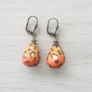 Puede incluir: Un par de pendientes con forma de lágrima con diseño de estampado de leopardo. Los pendientes presentan un degradado de naranja y amarillo con manchas negras y detalles dorados. Los ganchos para las orejas son de color bronce oscuro.