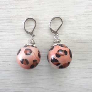 Puede incluir: Un par de pendientes redondos con diseño de estampado de leopardo. Los pendientes tienen una base de color melocotón con manchas marrones oscuras y negras. Cuentan con cierres de palanca plateados y detalles decorativos plateados.