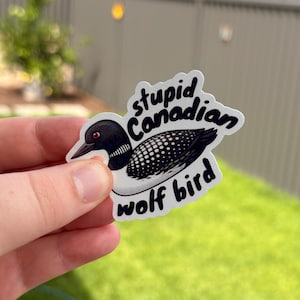May include: A sticker with a cartoon loon illustration and the words "stupid Canadian wolf bird". The bird is depicted in black, white, and grey, with a red eye, against a white background.