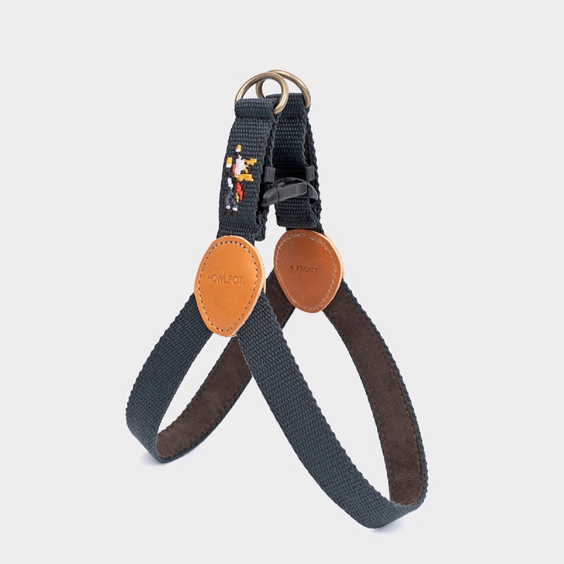 May include: A navy blue dog harness with brown leather accents and a pixelated design. The harness features a metal ring, a black plastic buckle, and the brand name "HOWLPOT" on a leather patch.