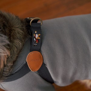 May include: A close-up of a dog wearing a gray harness with a black strap and a brown leather tag. The tag is stamped with the word "HOWLPOT." The harness has a small embroidered design and a brass ring.