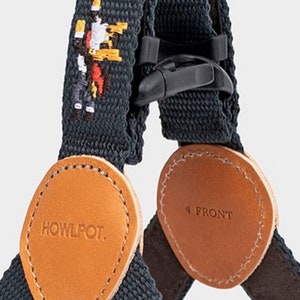 May include: Close-up of a dark gray dog harness with brown leather accents. The harness features a black plastic buckle and a stitched pixelated graphic. The leather patches are oval-shaped, with the text "HOWLPOT." and "FRONT" printed on them.