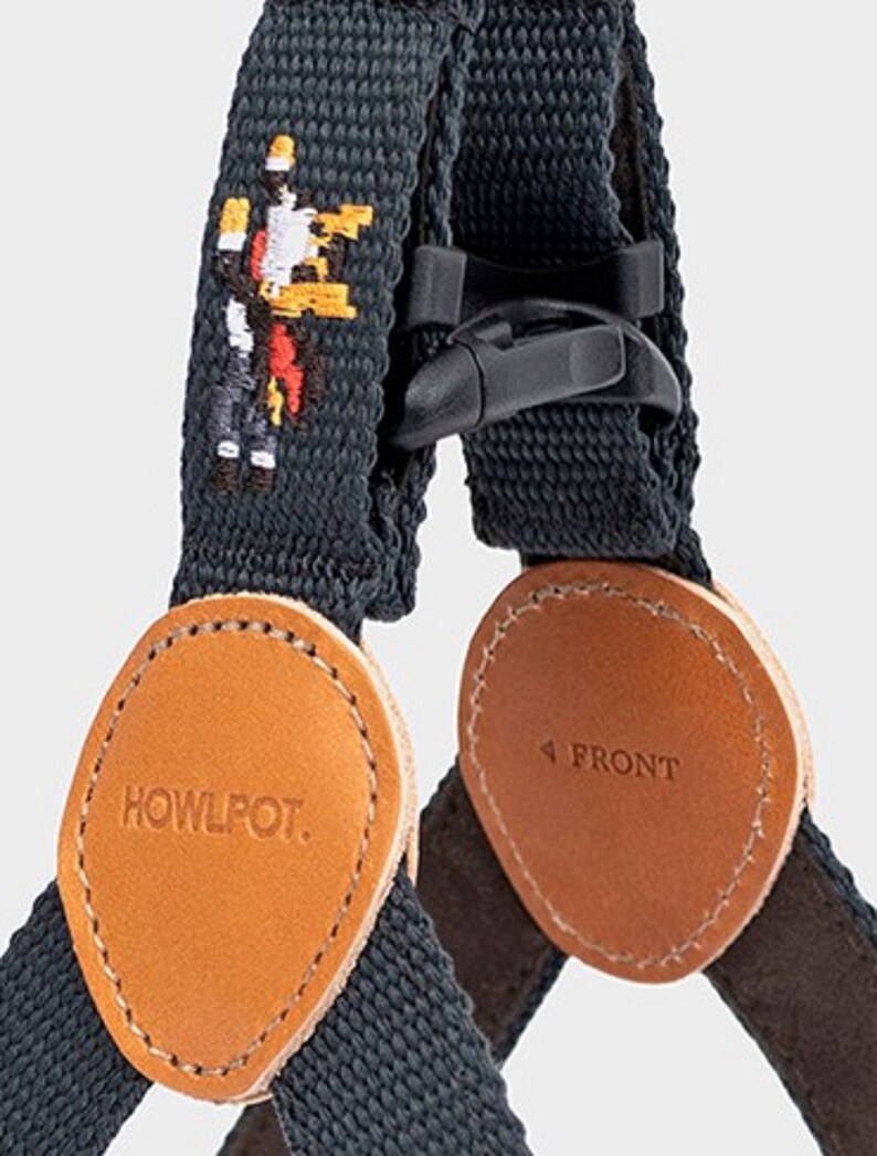 May include: Close-up of a dark gray dog harness with brown leather accents. The harness features a black plastic buckle and a stitched pixelated graphic. The leather patches are oval-shaped, with the text "HOWLPOT." and "FRONT" printed on them.