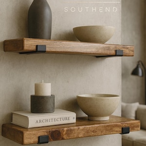 May include: Two horizontal wooden floating shelves mounted on a wall with black metal brackets. The top shelf holds a dark gray vase and a beige bowl. The bottom shelf holds a white candle in a gray holder, a book titled ARCHITECTURE, and a beige bowl.