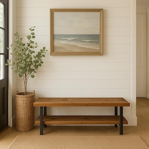 May include: A wooden bench with a lower shelf and black metal legs sits beneath a framed landscape painting of a beach scene. A tall woven basket holds a leafy plant, adding a touch of nature to the interior decor.