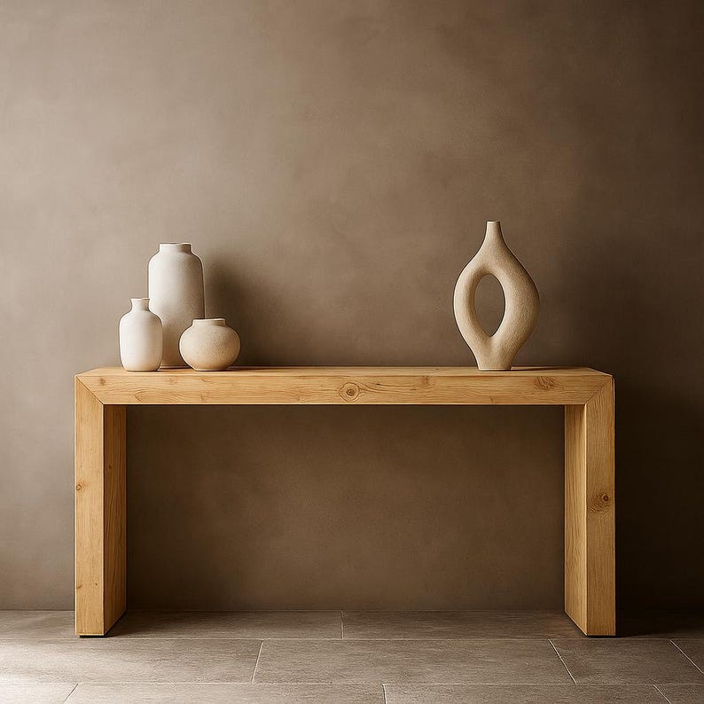 Puede incluir: Una mesa consola de madera clara con una superficie rectangular y dos patas verticales. En el lado izquierdo, hay tres jarrones de cer&aacute;mica de color blanco roto mate de diferentes alturas y formas. A la derecha, un jarr&oacute;n escultural.