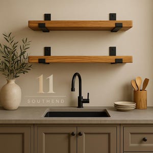 May include: Two horizontal wooden shelves with black metal brackets mounted on a wall above a kitchen sink. The sink features a black faucet. To the left is a beige vase with greenery, and to the right are wooden utensils and stacked plates.