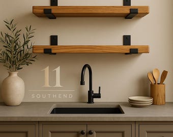 May include: Two horizontal wooden shelves with black metal brackets mounted on a wall above a kitchen sink. The sink features a black faucet. To the left is a beige vase with greenery, and to the right are wooden utensils and stacked plates.