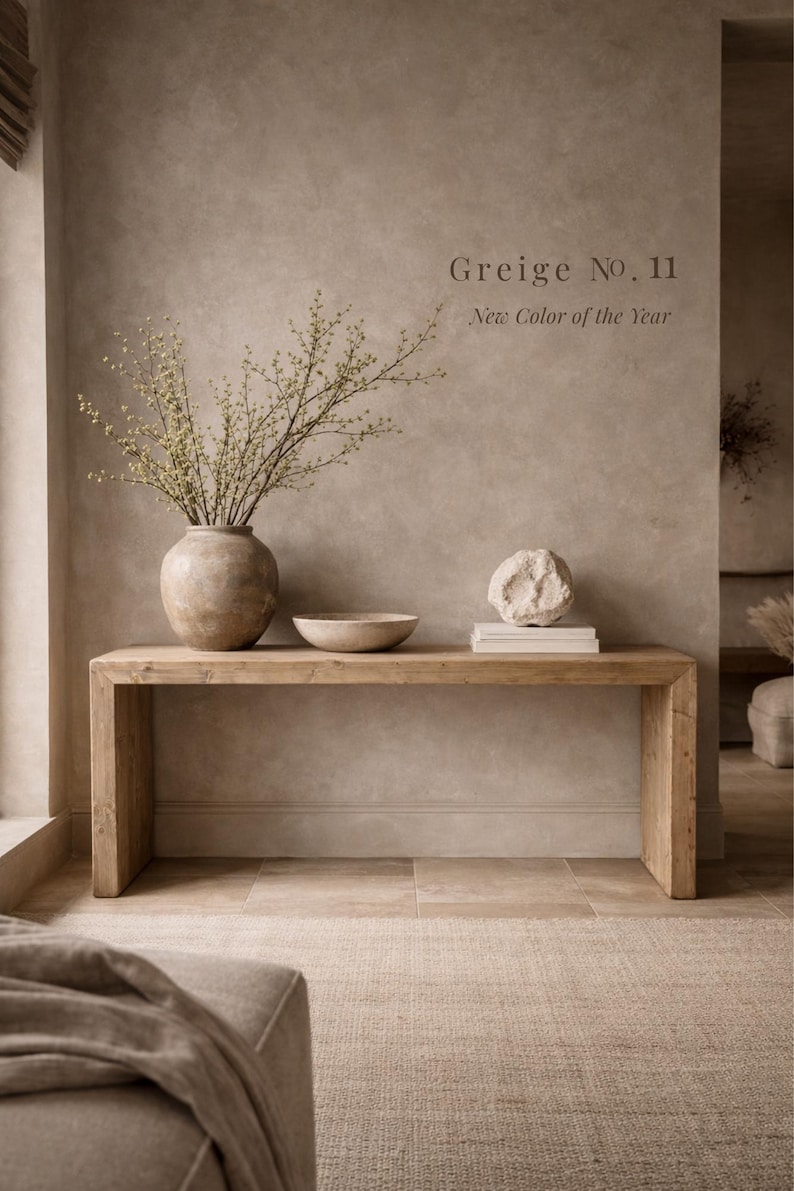 Puede incluir: Una mesa consola rectangular de madera con acabado natural, sobre la que hay un gran jarr&oacute;n de cer&aacute;mica con ramas verdes, un cuenco de cer&aacute;mica poco profundo y una piedra texturizada sobre libros blancos, con el texto Greige No. 11 New Color of the Year.