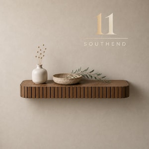May include: A rectangular wooden wall shelf with a vertical slatted front design. On the shelf sits a small, round, off-white ceramic vase containing dried stems and a shallow, textured, beige stone bowl with a green olive branch. Text reads 11 SOUTHEND.