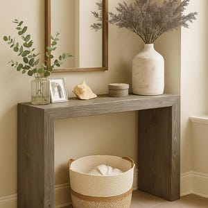 Puede incluir: Una consola de madera gris con marco rectangular y un espejo arqueado encima. Sobre la mesa hay un jarr&oacute;n de cristal estriado con eucalipto, un peque&ntilde;o marco de fotos, una concha, una caja redonda y un jarr&oacute;n blanco grande con follaje seco.