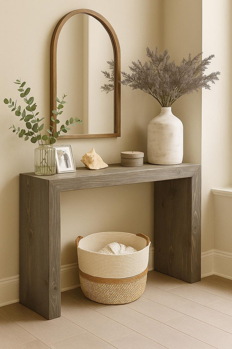 Puede incluir: Una consola de madera gris con marco rectangular y un espejo arqueado encima. Sobre la mesa hay un jarr&oacute;n de cristal estriado con eucalipto, un peque&ntilde;o marco de fotos, una concha, una caja redonda y un jarr&oacute;n blanco grande con follaje seco.