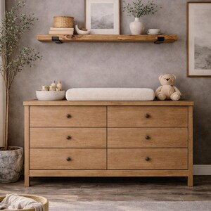 May include: A light wood dresser with six drawers, topped with a changing pad and a teddy bear. A wooden shelf above holds decor. The room has a neutral color palette, with a woven basket in the foreground.