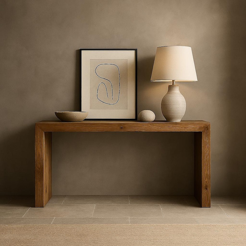 Puede incluir: Una mesa consola rectangular de madera con acabado de veta natural. Encima hay un cuenco de cer&aacute;mica beige, un dibujo lineal abstracto enmarcado en negro sobre fondo crema, una peque&ntilde;a piedra lisa y una l&aacute;mpara con base estriada y pantalla.