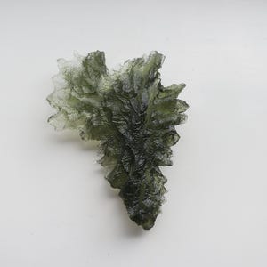 May include: A green, translucent, crystalline formation with a rough, textured surface. The crystal is shaped like a leaf and has a pointed tip.