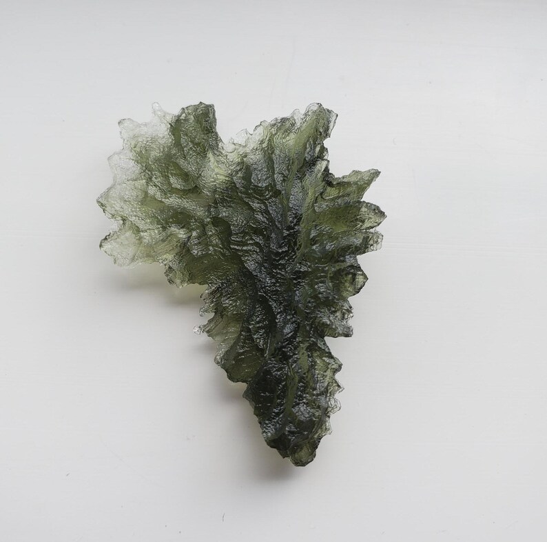 May include: A green, translucent, crystalline formation with a rough, textured surface. The crystal is shaped like a leaf and has a pointed tip.