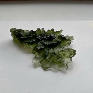 May include: A green, translucent, irregularly shaped piece of moldavite, a type of tektite, on a white surface.