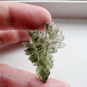 May include: A green, translucent, and irregularly shaped piece of Moldavite, a type of tektite, held in a hand.
