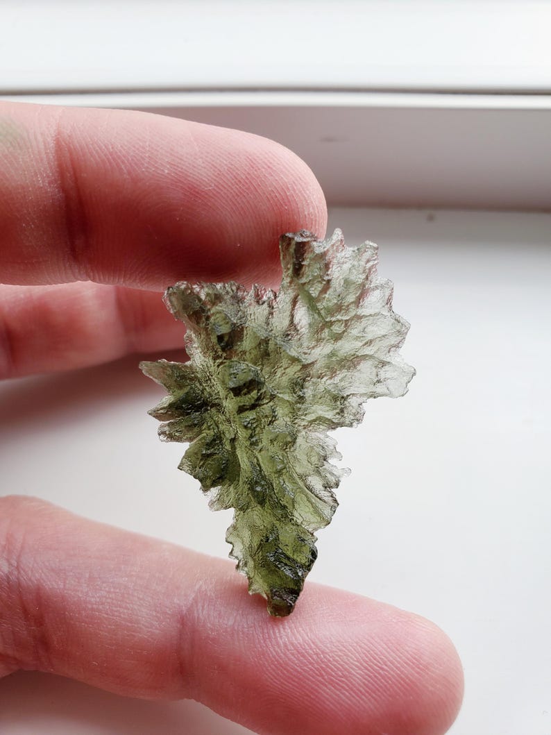 May include: A green, translucent, and irregularly shaped piece of Moldavite, a type of tektite, held in a hand.