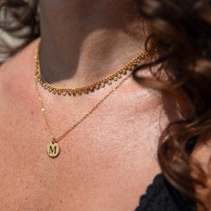 Peut inclure: Un collier en chaîne dorée avec un petit pendentif rond orné de la lettre "M". Le collier est superposé à une délicate chaîne dorée avec de petites perles rondes.