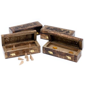 Puede incluir: Cuatro cajas de madera con tallas intrincadas y acentos de elefante dorados. Las cajas están abiertas y revelan pequeños discos redondos dorados. Las cajas están diseñadas para contener conos de incienso. Hay seis conos de incienso sobre una superficie blanca.
