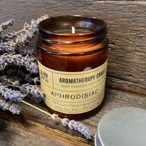 Peut inclure: Un bocal en verre brun contenant une bougie avec une étiquette blanche indiquant "Aromatherapy Candle, Pure Essential Oils + Soy Wax, Aphrodisiac, with 100% pure Ylang Ylang and Patchouli Essential Oil, 200g Soy Wax / 40h Burning Time".
