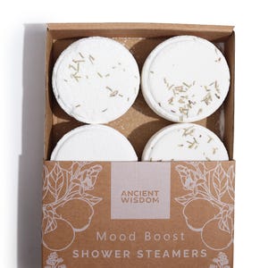Può includere: Una scatola di quattro pastiglie di vapore per doccia bianche su uno sfondo marrone. La scatola ha il testo "Ancient Wisdom" e "Mood Boost Shower Steamers".
