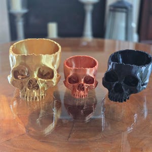 May include: Three skull-shaped decorative containers in gold, copper, and black. The containers are open at the top, with detailed facial features. They are displayed on a reflective surface, showcasing their unique design.