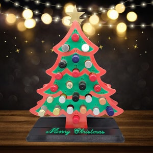 May include: A red and green Christmas tree advent calendar with 25 numbered compartments. Each compartment has a small, round, colorful lid. The tree is on a black base with the words "Merry Christmas" written on it.