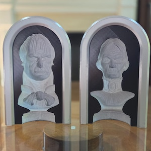 May include: Two decorative glass busts, each set within an arched frame. The busts depict the faces of two individuals, one male and one female, with detailed facial features. The frames have a black background and a silver border. The busts are displayed on a wooden surface.