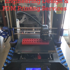 May include: A black 3D printer with a red filament spool holder and a blue and black textured base. The printer is displaying a screen with the text "8240/240° 2 14.60 90/90° 100% SD 90% 000:200 2MP PETG MK3S-4h138" and the word "ULTISTIK".