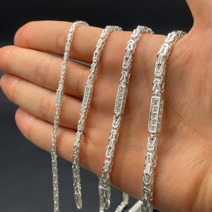 May include: Four silver chains with a box clasp, each with a different length. The chains are made of a woven pattern and have a small square of cubic zirconia stones on the clasp.