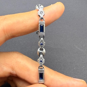 Puede incluir: Una pulsera de eslabones de metal plateado con piedras rectangulares de color azul oscuro que se alternan con segmentos plateados ornamentados con pequeños detalles azules. El diseño presenta patrones geométricos.