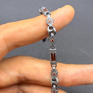 Peut inclure: Un bracelet en métal argenté composé de maillons rectangulaires sertis de pierres rouge foncé alternant avec des maillons circulaires texturés et ornés de petites pierres rondes rouge orangé, formant un design structuré et détaillé.