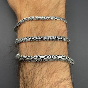May include: Three silver Byzantine chain bracelets are shown on a wrist. The bracelets feature a detailed, woven design. The bracelets are made of a shiny, metallic material. The background is a dark grey colour.