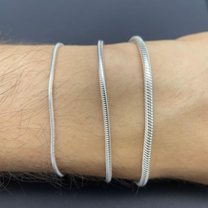 May include: Three silver snake chain bracelets of varying thicknesses are displayed on a person's wrist. The bracelets have a textured, flexible design, reflecting light. The background is a dark gray.