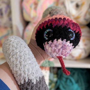 May include: A handmade crochet snake toy with a plush, textured body. The snake's head is black, pink, and purple with large, glittery eyes and a red tongue. The body transitions from gray to orange and blue.