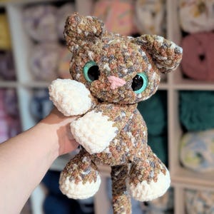 May include: A handmade crochet cat plush toy crafted from variegated brown, tan, and grey yarn. The cat features large, glossy teal eyes, a small pink stitched nose, and solid cream-colored paws. The texture is soft and plush throughout.