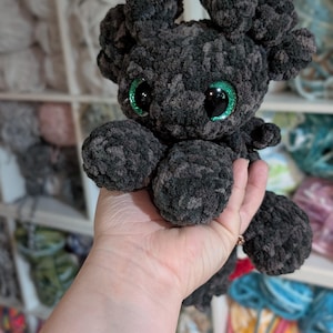 May include: A handmade, dark gray crocheted plush toy with large, glittery green eyes. The plush has a rounded body, four stubby limbs, and two small, ear-like protrusions on its head. The toy is held in a person's hand, with a background of yarn.