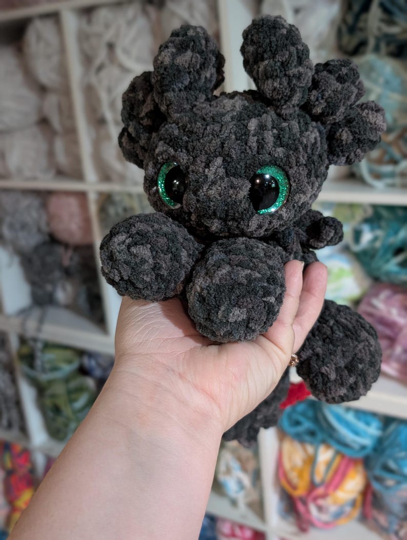 May include: A handmade, dark gray crocheted plush toy with large, glittery green eyes. The plush has a rounded body, four stubby limbs, and two small, ear-like protrusions on its head. The toy is held in a person's hand, with a background of yarn.