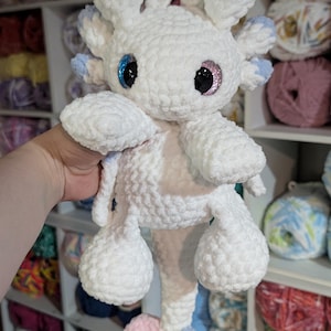 May include: A handmade white crochet axolotl plushie with blue and pink accents. The plushie has large, glittery eyes and a fluffy texture. The axolotl is being held in front of a shelf filled with yarn.