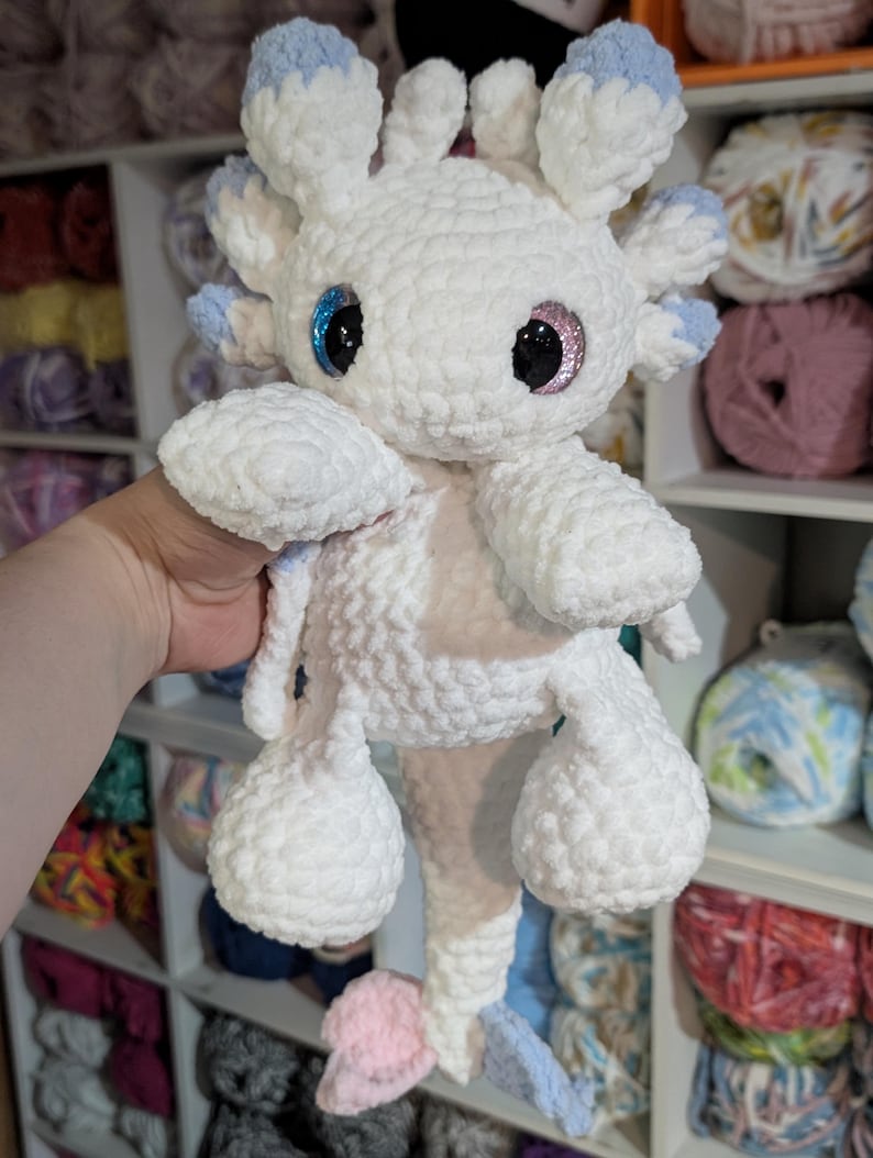 May include: A handmade white crochet axolotl plushie with blue and pink accents. The plushie has large, glittery eyes and a fluffy texture. The axolotl is being held in front of a shelf filled with yarn.