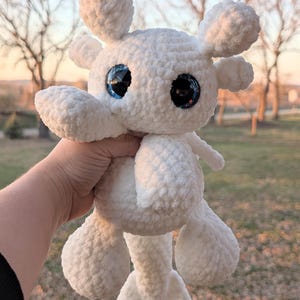 May include: A handmade white crochet stuffed animal with large, reflective blue and black eyes. The plush toy has a rounded body, four limbs, and two small, rounded ears. The toy is held against a blurred outdoor background.