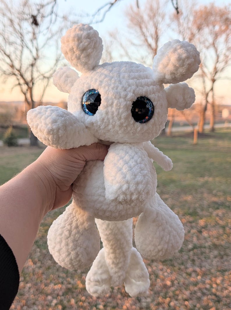 May include: A handmade white crochet stuffed animal with large, reflective blue and black eyes. The plush toy has a rounded body, four limbs, and two small, rounded ears. The toy is held against a blurred outdoor background.
