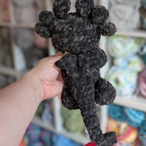 May include: A handmade, dark gray crocheted dragon plushie with a red tail. The dragon has a textured, plush appearance, with a head featuring multiple rounded protrusions. The tail is a vibrant red, contrasting with the gray body. The plushie is held in a hand.